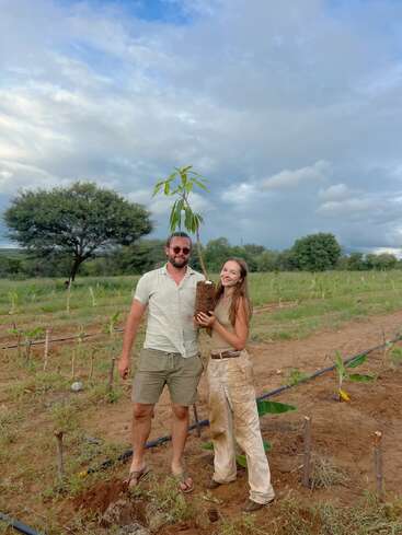 Um homem e uma mulher estão sorrindo em um campo, segurando uma árvore jovem, preparando-se para plantá-la. O plano de fundo mostra árvores verdes e um céu nublado.