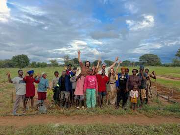 A joyful group of diverse people stand together outdoors, smiling and raising their hands, celebrating teamwork on a farm or garden with green fields and cloudy skies.