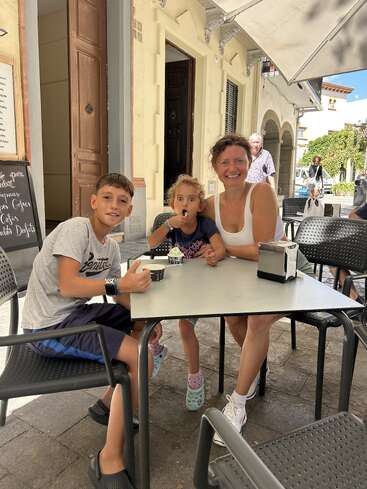 A smiling woman and two children sit at an outdoor café table, enjoying dessert. Sunlight, relaxed atmosphere, old building in the background, creating a pleasant summer scene.