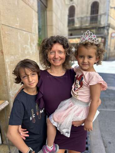 Das Bild zeigt eine Frau, die mit zwei Kindern, einem Jungen und einem Mädchen, auf einer Straße in der Stadt posiert und einen zärtlichen Moment zwischen den dreien zeigt.