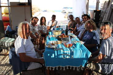 Un grupo de personas se reúne en torno a una mesa cubierta con un mantel azul, compartiendo comida y bebida, sonriendo y disfrutando de la compañía de los demás en un ambiente soleado y relajado.
