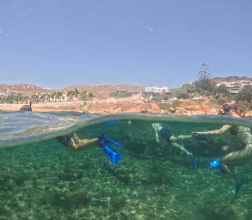 Esta imagen muestra a unos buceadores bajo el agua con aletas azules, un cielo despejado por encima, un paisaje costero y colinas al fondo, captadas en la superficie del agua.