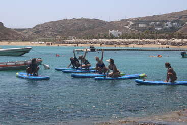 Un grupo de personas practica yoga en tablas de paddle surf en aguas tranquilas de color turquesa cerca de una playa, con montañas, sombrillas y un complejo turístico visibles al fondo.