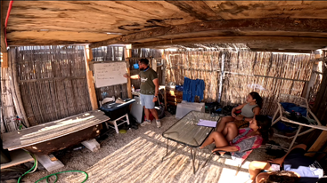Un grupo se reúne en una rústica aula al aire libre con paredes de bambú. Una persona expone en una pizarra, mientras otras tres escuchan sentadas alrededor de una mesa de cristal.