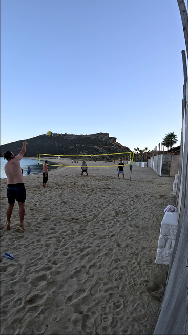 Cuatro personas juegan al vóley-playa en una playa de arena durante la puesta de sol. La red está colocada, el mar y una montaña proporcionan un pintoresco telón de fondo natural.
