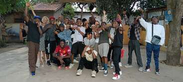 A lively group of young people posing together outdoors, many smiling and making playful hand gestures. They express joy, unity, and friendship in a casual setting.