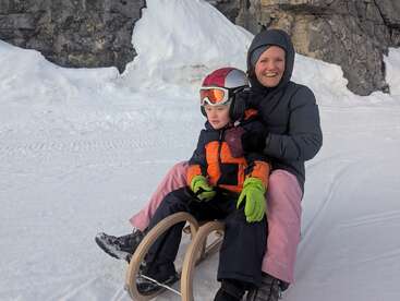 Uma mulher e uma criança sentam-se juntas em um trenó de madeira em montanhas nevadas. Ambos estão vestidos com roupas quentes; a criança usa um capacete e óculos de proteção, pronta para se divertir.