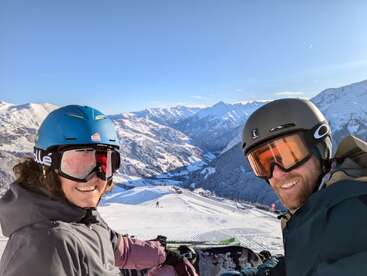 Duas pessoas usando capacetes de esqui e óculos de proteção sorriem para a câmera em uma montanha nevada, cercada por belos picos ensolarados cobertos de neve e um céu azul claro.