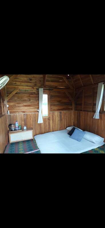 This image shows a cozy wooden cabin interior with a mattress on the floor, pillows, a nightstand, small windows, curtains, a fan, and kettle.