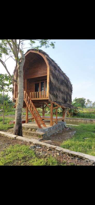 A small wooden treehouse stands on stilts, featuring a thatched roof, balcony, and staircase, surrounded by lush greenery and a peaceful rural landscape.