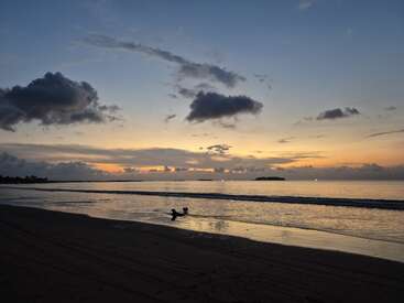 A serene beach at dusk, sky painted with soft orange and blue hues. Silhouetted figures wade in gentle waves, clouds drifting above the peaceful, reflective water.