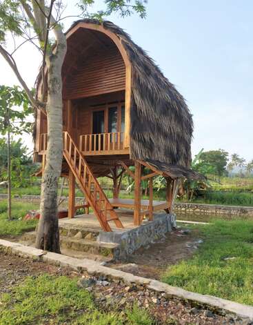 A small wooden treehouse stands on stilts, featuring a thatched roof, balcony, and staircase, surrounded by lush greenery and a peaceful rural landscape.