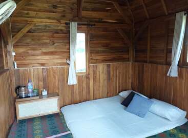 This image shows a cozy wooden cabin interior with a mattress on the floor, pillows, a nightstand, small windows, curtains, a fan, and kettle.