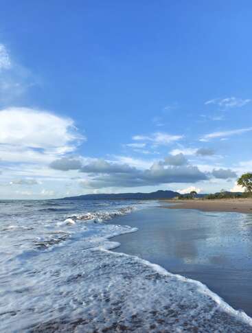 Waves gently reach the sandy shore under a bright blue sky. White clouds drift above, while distant mountains and trees complete the peaceful coastal scenery.