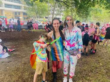 Three people stand together outdoors at a colorful event, wearing rainbow and tie-dye clothes. The child holds a toy bow; people celebrate in the vibrant background.