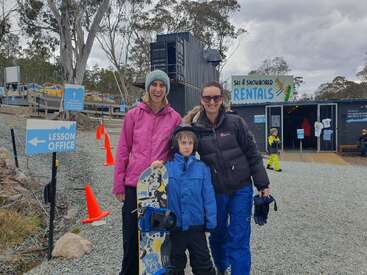 Three people, two women and a boy in winter clothing, pose outside a ski and snowboard rental shop. Orange cones and signs for lessons are visible.