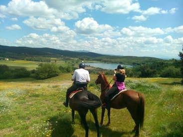 Das Bild zeigt zwei Menschen, die auf Pferden in einem malerischen Feld mit Wildblumen reiten, mit Blick auf einen ruhigen See und ferne Berge unter einem blauen Himmel mit weißen Wolken.