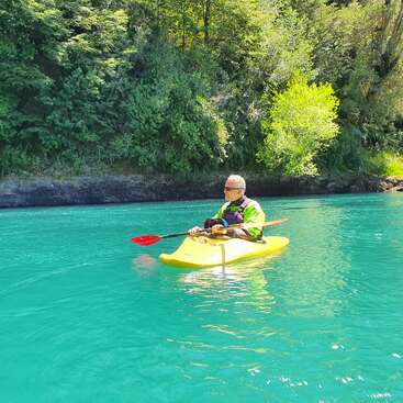 The image depicts an elderly man kayaking on a serene body of water, surrounded by lush greenery and trees, with a vibrant yellow kayak and a red paddle.