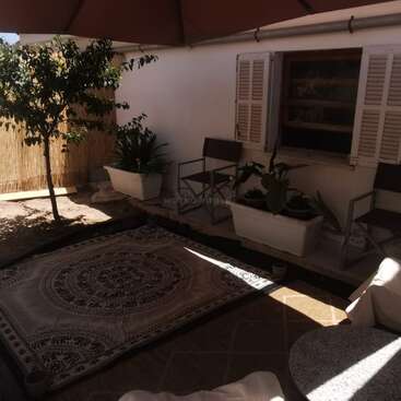 This cozy outdoor patio features a patterned rug, shaded seating with chairs, potted plants, a tree, bamboo fencing, and an umbrella, creating a relaxing atmosphere.