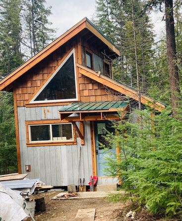 Nichée au cœur d'une forêt, cette petite cabane rustique se distingue par une fenêtre triangulaire unique, un revêtement en bois et en métal, un toit végétalisé et une porte bleue qui respire la chaleur.