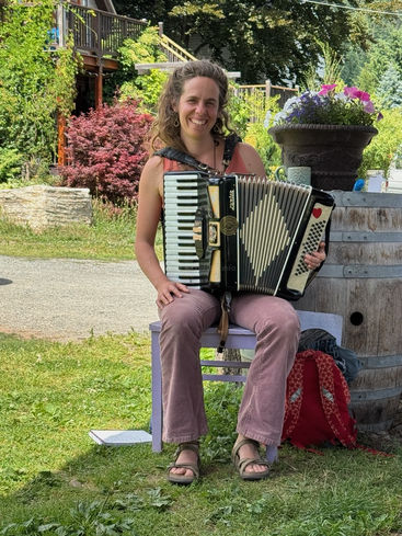 A woman is sitting outside on a small purple bench, smiling, playing an accordion. She is surrounded by plants, flowers, and summertime greenery.