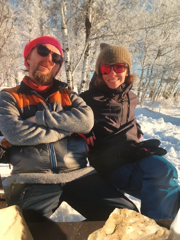 A man and woman sit together in snowy woods, both wearing sunglasses, winter hats, and jackets, smiling and enjoying a sunny winter day outdoors.