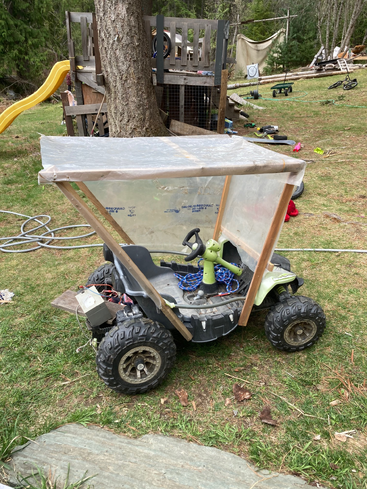 A toy car with a homemade canopy made of plastic and wood sits on grass. In the background, there’s a playground structure, yellow slide, and outdoor toys.