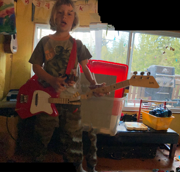 A young child with shoulder-length hair stands indoors playing a red electric guitar. The room is cluttered, and there appears to be a digital glitch effect.