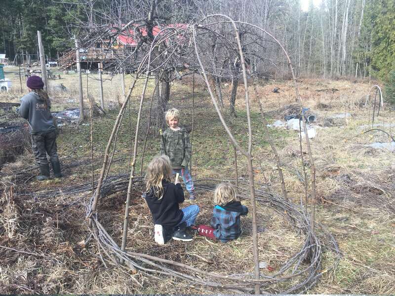 Three children play inside a homemade stick structure outdoors. The setting appears rural, with leafless trees and a rustic house in the background on a cloudy day.