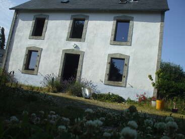 This is a tilted photo of a white two-story house with six windows, a front door, surrounding plants, and a grassy garden in the foreground.