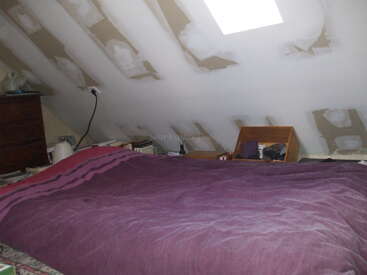 This image shows an attic bedroom with unfinished drywall, a purple bedspread, wooden furniture, a skylight, and various items scattered around, creating a cozy yet cluttered atmosphere.