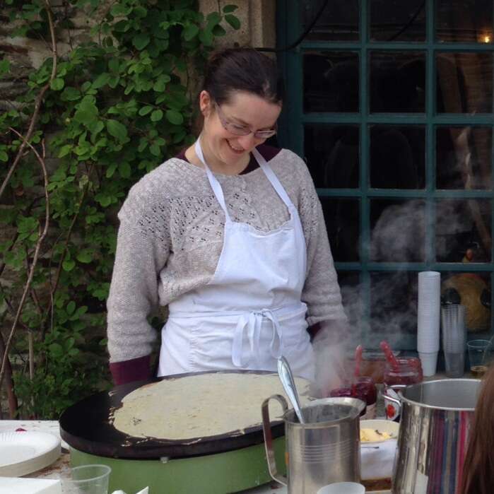 A woman wearing glasses and a white apron smiles while making a large crepe on a griddle outdoors, surrounded by cooking utensils, steaming pots, and greenery.