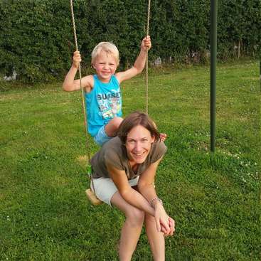 Das Bild zeigt eine Frau und einen kleinen Jungen auf einer Schaukel, vor dem Hintergrund einer saftigen Wiese mit Bäumen in der Ferne.
