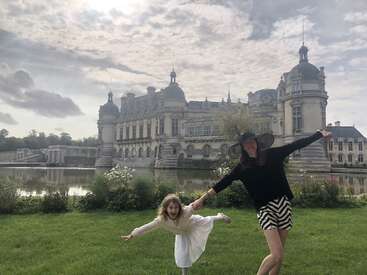 Eine Frau und ein junges Mädchen posieren freudig auf grünem Gras und halten sich mit ausgestreckten Armen an den Händen, vor einem großen europäischen Schloss mit Wassergraben.