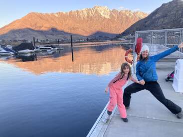 Eine Frau und ein Kind posieren spielerisch auf einem Steg an einem ruhigen See, mit schneebedeckten Bergen und Booten im Hintergrund bei Sonnenuntergang. Schöne Spiegelungen erscheinen.
