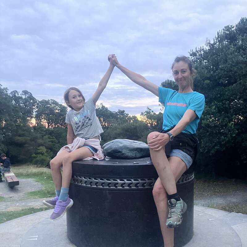 Eine Frau und ein Mädchen sitzen auf einer großen schwarzen Skulptur im Freien, halten die Hände in die Luft und lächeln, umgeben von Bäumen unter einem bewölkten Abendhimmel.