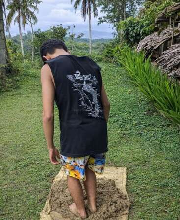 A person stands barefoot in mud on a mat, wearing a black sleeveless shirt and colorful shorts. Surrounded by lush greenery, they seem relaxed and peaceful.