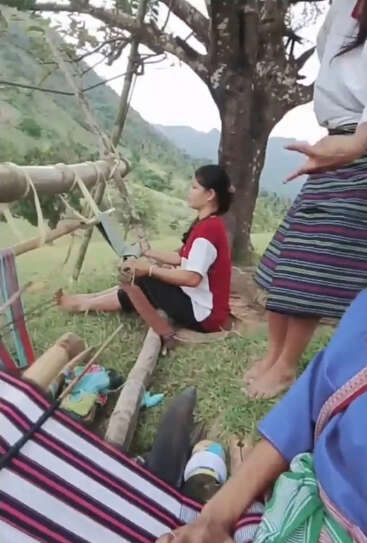 Three women, dressed in traditional clothing, gather outdoors under a tree on a hillside, practicing indigenous weaving techniques amidst nature, preserving their cultural heritage and skills.