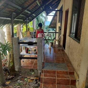 A cozy private balcony with tiled floor, pantry kitchen, and overhead roof. A person stands facing lush greenery, enjoying the ocean view and gentle breeze. Peaceful atmosphere.