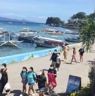 People stroll along a sunny baywalk in Sabang, beside colorful boats anchored on clear blue water. Lush greenery and vibrant street life create a lively seaside atmosphere.