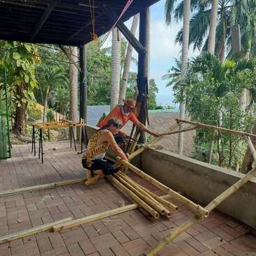 Two people collaboratively constructing a bamboo structure on a brick patio surrounded by lush tropical trees, focused and engaged in their hands-on project under a covered area.