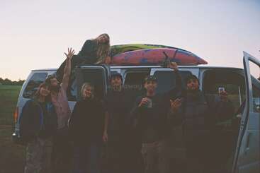 A group of seven friends stand in front of a white van at sunset, posing happily with drinks, while one person lounges on top with kayaks.