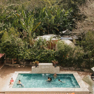 Ein paar Leute chillen an einem Pool, der von viel Grün und tropischen Pflanzen umgeben ist. In der Nähe sieht man ein Haus mit rotem Dach und Terrasse.