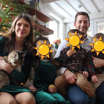 Une famille souriante est assise près d'un sapin de Noël. La femme tient un chat dans ses bras. Les visages des enfants sont recouverts d'émojis représentant des soleils. Tout le monde semble joyeux et festif.