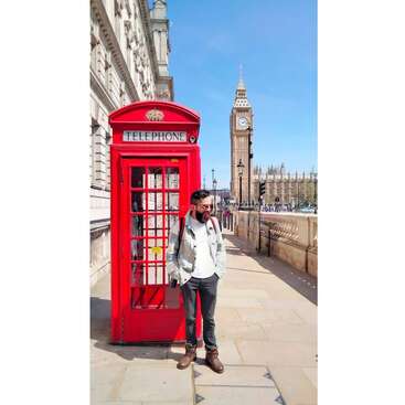 The image depicts a man standing beside a red telephone booth on a sidewalk, with the iconic Big Ben clock tower visible in the background.