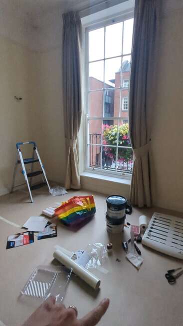 The image depicts a room in the process of renovation, featuring a window with curtains, a ladder, and scattered tools and materials on the floor.