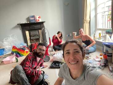 The image shows four people sitting on the floor of a room filled with cleaning supplies and tools, surrounded by a cluttered space.