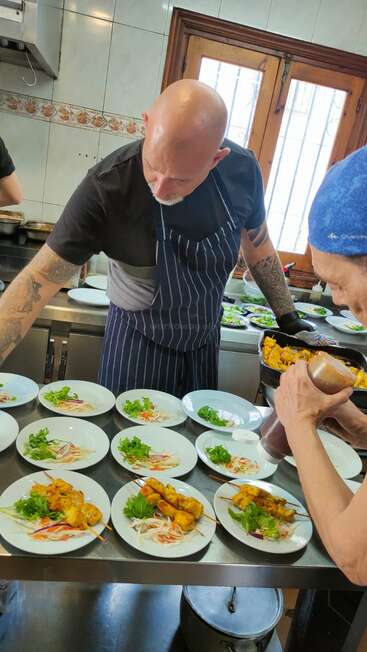 Dans une cuisine professionnelle, deux chefs préparent des brochettes de viande grillée accompagnées de légumes frais. Ils préparent plusieurs plats identiques pour le service ou pour un événement.