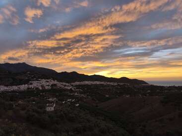 Un coucher de soleil époustouflant peint le ciel de teintes dorées et violettes sur un paysage montagneux tentaculaire, un village blanc niché paisiblement au milieu de collines vertes foncées.