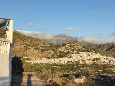 Un village blanc pittoresque est niché au milieu de collines et de montagnes verdoyantes, sous un ciel bleu clair, avec des nuages doux et un soleil chaud qui illuminent le paysage paisible.